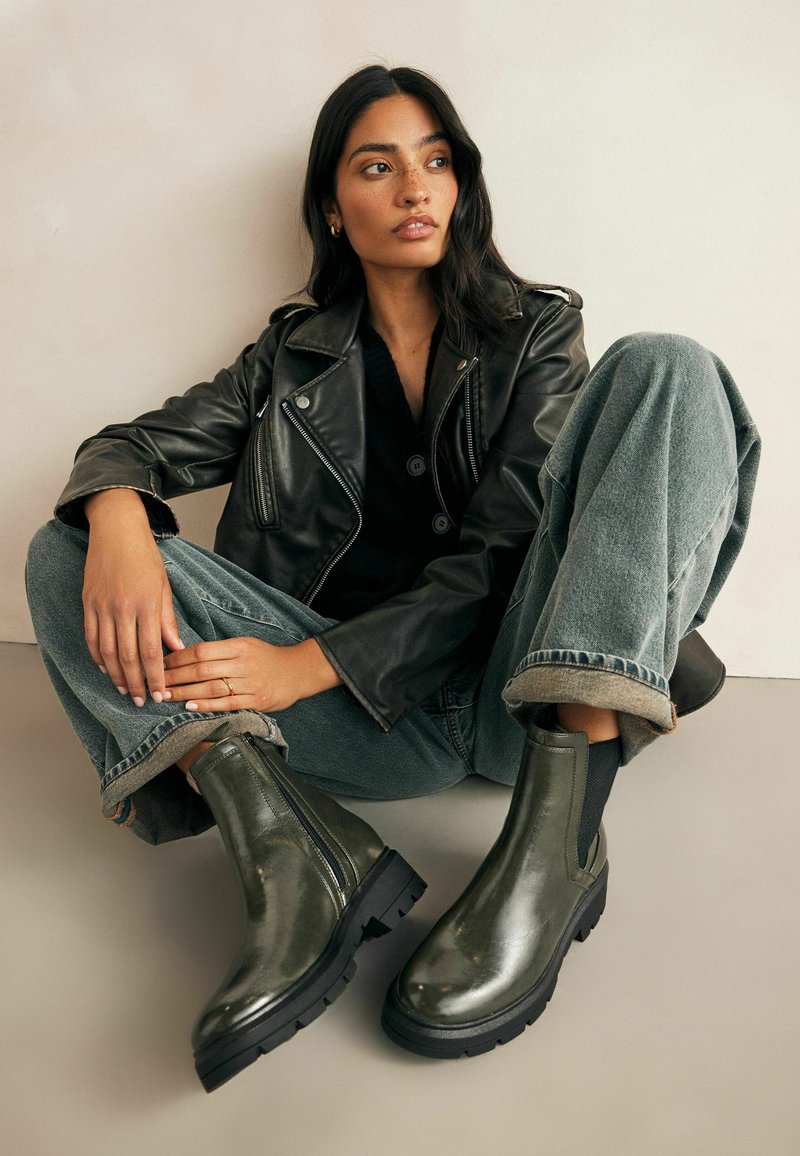 Botas de cuero verde oliva hasta el tobillo con cremallera lateral, suela negra gruesa y textura suave, combinadas con unos vaqueros relajados y una chaqueta de cuero.
