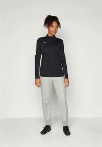 Zwarte sportieve zip-up top met lange mouwen en logo, gecombineerd met grijze sweatpants met boorden en zwarte sportschoenen.