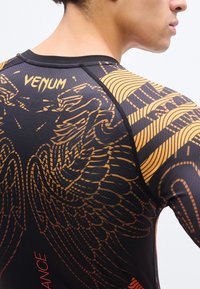 Κοντινό πλάνο ενός ατόμου που φοράει αθλητική μπλούζα Venum μαύρου και πορτοκαλί χρώματος με φτερωτό σχέδιο στην πλάτη και κόκκινο κείμενο στην πλευρά.