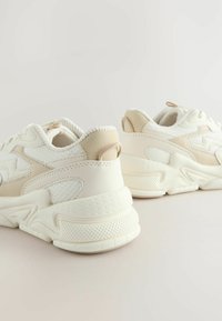 Des baskets blanches et beiges en matériaux mesh et cuir, avec une semelle texturée, une forme arrondie et des accents de design minimalistes.