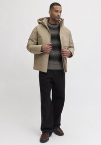 Beige isolierte Jacke mit Reißverschluss und Kapuze, getragen über einem dunkel gestreiften Pullover und kombiniert mit dunkler Hose und braunen Schuhen.