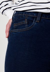 Jean bleu foncé en denim, avec une poche avant, des détails de couture et une texture lisse. Porté avec une chemise rayée noire et blanche.