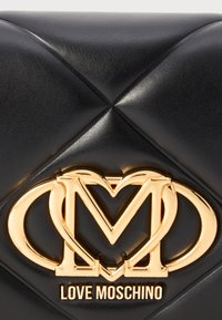 Schwarze Lederhandtasche mit einem gesteppten Design, einem Logo aus goldfarbigem Metall und einer kompakten Form. Dekorative Nähte akzentuieren die Oberfläche.