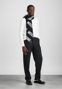 Μαύρο κασκόλ με λευκό λογότυπο "Moschino", φορεμένο πάνω από ένα λευκό πουλόβερ, συνδυασμένο με μαύρα παντελόνια και μαύρα παπούτσια, σε ανοιχτό γκρι φόντο.