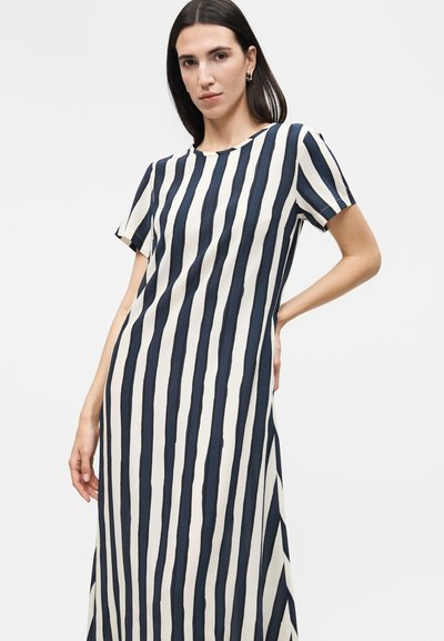 Γυναίκα με ίσια σκούρα μαλλιά που φοράει φόρεμα με κοντά μανίκια και κατακόρυφες ασπρόμαυρες ρίγες μέχρι τους αστραγάλους.
