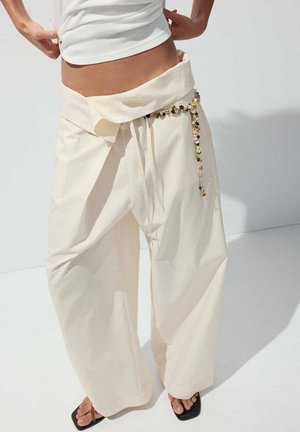 Persona que lleva pantalones anchos color crema, sueltos, con detalle de envoltura y cinturón con cuentas, combinados con sandalias negras de punta cuadrada.