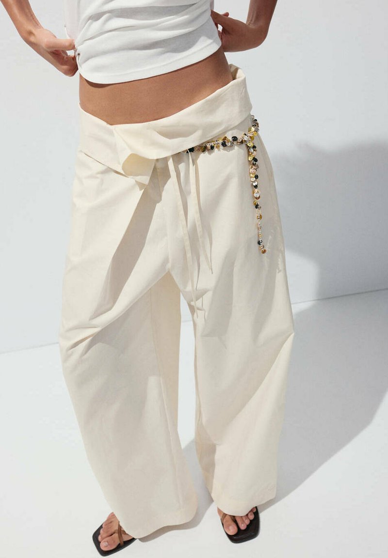 Personne portant un pantalon large crème ample avec un détail noué et une ceinture perlée, associé à des sandales noires à bout carré.