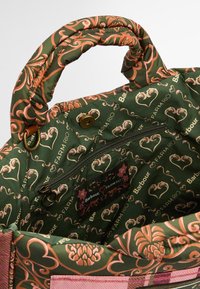 Gestepptes grünes Täschchen mit floralen Mustern in Rosa und Gold. Verfügt über einen dicken Griff und eine Reißverschlusstasche innen, mit sichtbarem Branding.