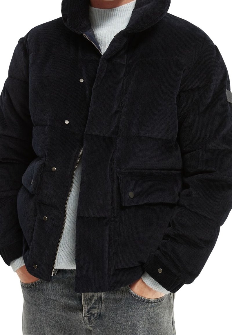 Giacca puffer in corduroy blu navy con colletto alto, zip frontale, chiusure a scatto e due tasche laterali con polsini a coste.