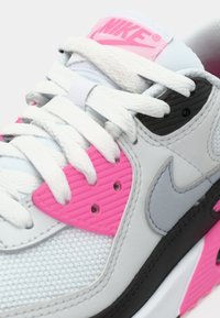 Nike sportbačiai su šviesiai pilku viršumi, rožinėmis akcentais ir tekstūruotu pilku panele. Balti raišteliai ir juodi detalės užbaigia dizainą.