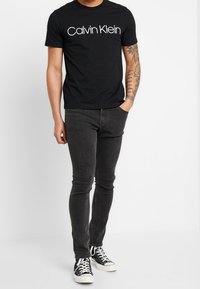 Černé tričko s bílým logem "Calvin Klein", spojené s tmavě šedými skinny džínami a černými teniskami, s jednoduchým designem a neformálním střihem.