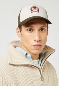 Jonge man in een beige gebreid trui met rits, lichtgroene geruite blouse en een beige en groene pet met een boomlogo.