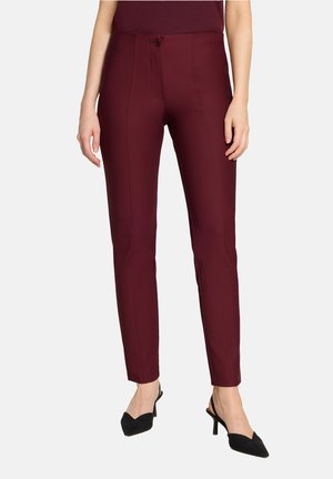 Pantalon slim fit bordeaux porté avec des talons noirs pointus à bride arrière, présenté de la taille aux pieds sur un fond uni.