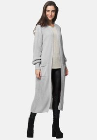 Lång, grå, stickad cardigan med två framfickor, öppen fram och textil med struktur. Bärs över en beige topp och svarta byxor.