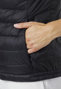Schwarze Steppweste mit gestepptem Muster, seitlichem Reißverschlussfach und elastischem Saum. Nahaufnahme einer Hand, die auf der Tasche ruht.