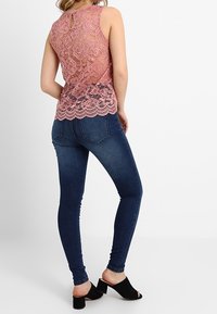 Růžový krajkový top s květinovým vzorem, vroubkovaným lemem a zadním detailem ve tvaru klíčové dírky, nošený s tmavě modrými skinny džíny a černými nazouváky s otevřenou špičkou.