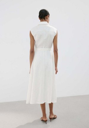 Femme debout pieds nus, dos à la caméra, portant une robe midi blanche sans manches dans une pièce claire et simple.