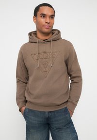 Hombre con sudadera marrón de Guess con logo y jeans de mezclilla azules, con las manos en los bolsillos, mirando ligeramente hacia la derecha contra un fondo blanco liso.