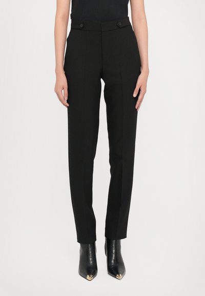 Schwarze, maßgeschneiderte Hose mit hoher Taille, zwei vorderen Knöpfen, Seitentaschen und schlanker Passform. Kombiniert mit spitzen schwarzen Stiefeletten.