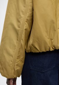 Zbliżenie na osobę noszącą musztardowo-żółtą pikowaną kurtkę z elastycznym obszyciem i niebieskie dżinsy, na których widać tylną kieszeń.