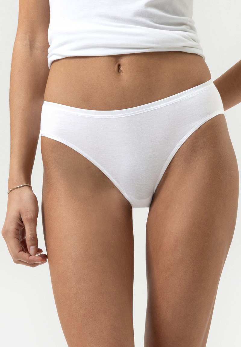 Frau in weißen Baumwollslips und einem weißen Shirt, von der Taille bis zur Mitte des Oberschenkels gezeigt, mit neutralem Hintergrund.