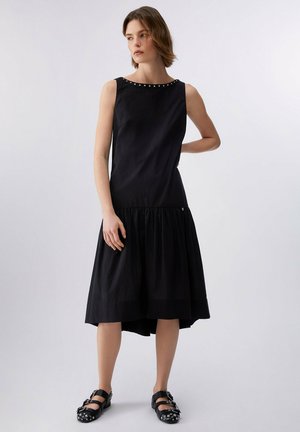 Kobieta ubrana w czarną sukienkę bez rękawów z nitowanymi detalami przy dekolcie oraz czarne nitowane buty typu Mary Jane, stojąca na jednolitym tle.