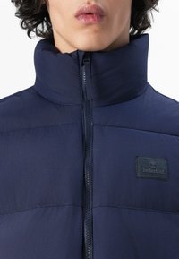 Marineblauwe pufferjack met een hoge kraag, ritssluiting en een Timberland-patch. Beschikt over een gladde textuur en gestikte panelen voor isolatie.