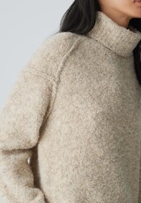 Beige, strukturierter Pullover mit hohem Stehkragen und überschnittenen Schultern; aus gemütlichem, weichem Material mit einem dezenten Muster gefertigt.