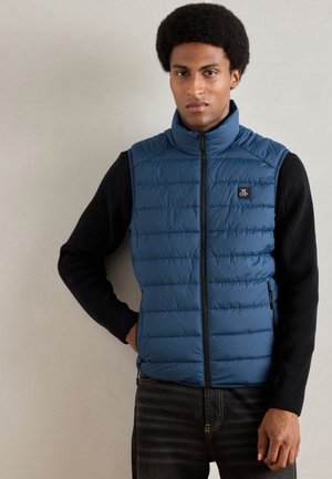 Blå puffervest med vertikal quiltet design, høj krave og lynlåslukning. Sammen med en sort langærmet top og mørke jeans.