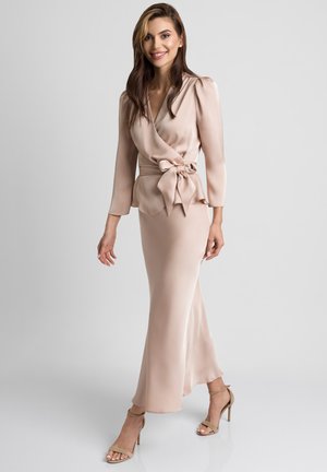 Uśmiechnięta kobieta idąca w beżowej satynowej bluzce z dużą kokardą i dopasowanej długiej spódnicy oraz sandałach na wysokim obcasie na szarym tle.