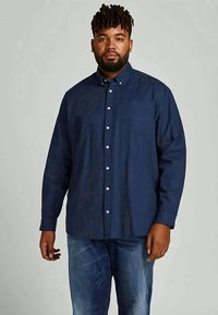 Homem com barba e dreads, vestindo uma camisa azul-marinho de botão e jeans azuis, de pé contra um fundo cinza simples.