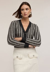 Cárdigan de rayas blancas y negras con un profundo escote en V, que cuenta con un cierre de un solo botón y puños acanalados, combinado con una falda texturizada color crema.