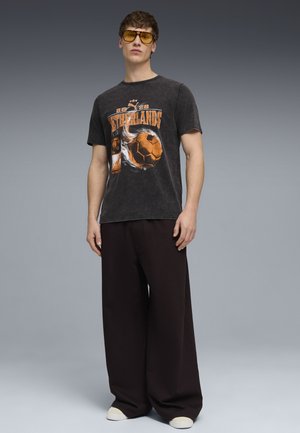 Jeune homme portant des lunettes de soleil teintées jaunes, un t-shirt noir délavé avec un motif des Pays-Bas, un pantalon large noir et des baskets blanches sur fond gris.