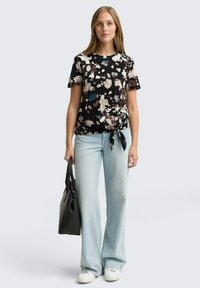 Donna in jeans azzurri chiari e maglietta nera a maniche corte con un motivo, che tiene una borsa nera e indossa sneakers bianche su uno sfondo semplice.