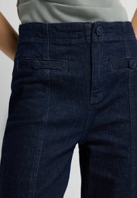 Tmavě modré džíny z denimu s hladkou texturou, vysokým pasem, dvěma předními kapsami a zapínáním na knoflík. Obsahují jemné švy podél švů.