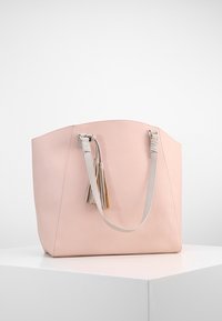 Sac cabas en cuir rose doux avec doubles anses d'épaule et franges décoratives, présenté sur un piédestal blanc sur un fond uni.