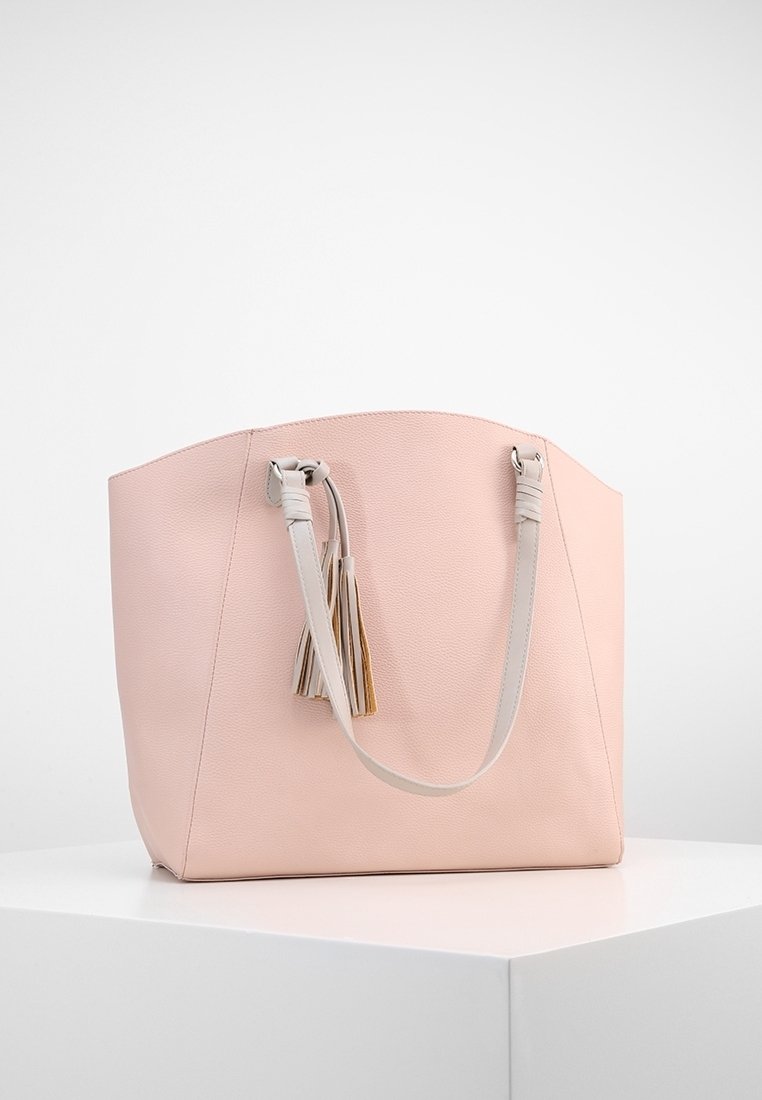 Sac cabas en cuir rose doux avec doubles anses d'épaule et franges décoratives, présenté sur un piédestal blanc sur un fond uni.