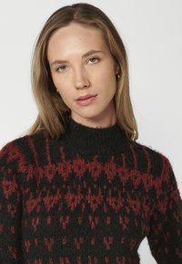 Pull noir et rouge à motif, avec un col montant, en tricot texturé, présentant des designs géométriques sur l'ensemble du tissu.