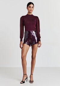 Top in magenta a maglia con maniche a palloncino abbinato a pantaloni corti lucidi in paillettes viola, completato da tacchi alti neri con cinturini.