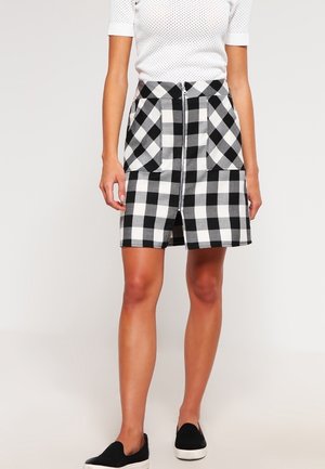 Nainen pukeutuneena mustavalkoruudulliseen hameeseen, jossa on edessä vetoketju ja kaksi taskua, yhdistettynä valkoiseen lyhythihaiseen toppiin ja mustiin slip-on-kenkiin.