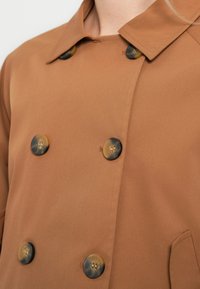 Εικόνα κοντινού πλάνου ενός καφέ διπλοκουμπωτού παλτό με έξι μεγάλα μαρμάρινα κουμπιά και μυτερό γιακά.