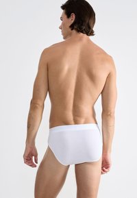 Mann mit schulterlangen Haaren, der schlichte weiße Unterhosen trägt, mit dem Rücken zur Kamera stehend vor einem neutralen Hintergrund.
