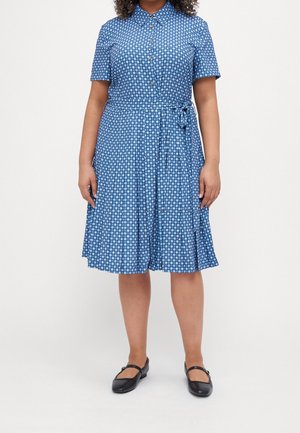 Femme portant une robe bleue à motifs à manches courtes avec boutons et ceinture nouée, associée à des chaussures Mary Jane noires, debout sur un fond blanc.
