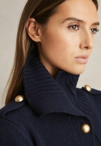 Pulover tricotat bleumarin cu guler înalt, având butoane mari aurii și o textură cusută. Prim-plan al zonei gulerului.