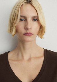 Una modella con capelli corti e biondi indossa una maglietta marrone a scollo a V. Il tessuto appare liscio, con una vestibilità comoda che valorizza il décolleté.