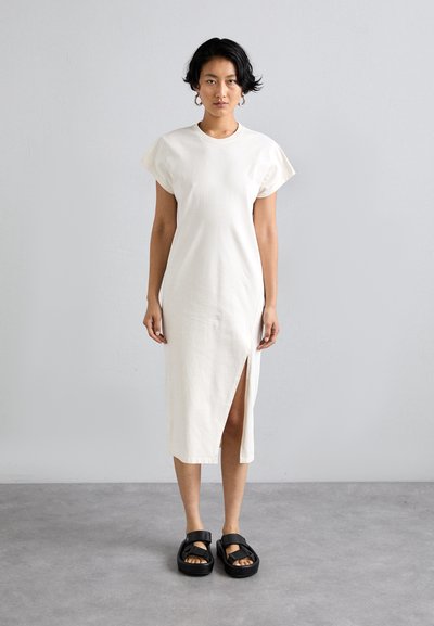 Cremefarbenes, knielanges Kleid mit kurzen Ärmeln und einem seitlichen Schlitz; kombiniert mit schwarzen Sandalen, die eine dicke Sohle und ein Riemendesign aufweisen.