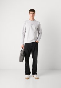 Felpa grigia con scollatura rotonda e piccolo logo, abbinata a jeans scuri e sneakers bianche. Tiene in mano una borsa grigia con manici.