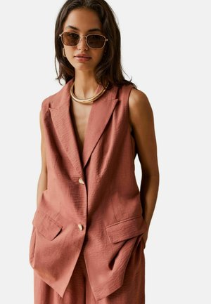 Mujer con blazer sin mangas de gran tamaño color rosa con bolsillos, pantalones a juego, collar dorado, pendientes de aro y gafas de sol tintadas.