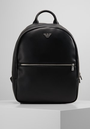 Sac à dos en cuir noir avec une poche zippée à l'avant, une poignée supérieure arrondie et un petit logo argenté près du centre supérieur.