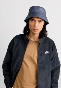 Nike klobúk typu bucket v tmavošedej farbe; svetlohnedé tričko pod čiernou kordurovouv šušťákovkou s bielym logom. Hladká textúra a jednoduchý dizajn.